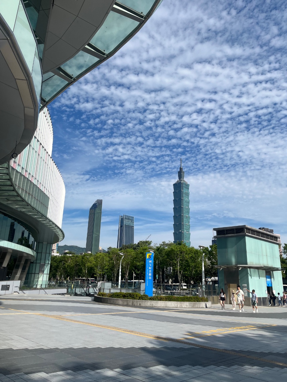 晴天下的台北101，前景為弧形玻璃建築與空曠廣場，幾名行人走過。