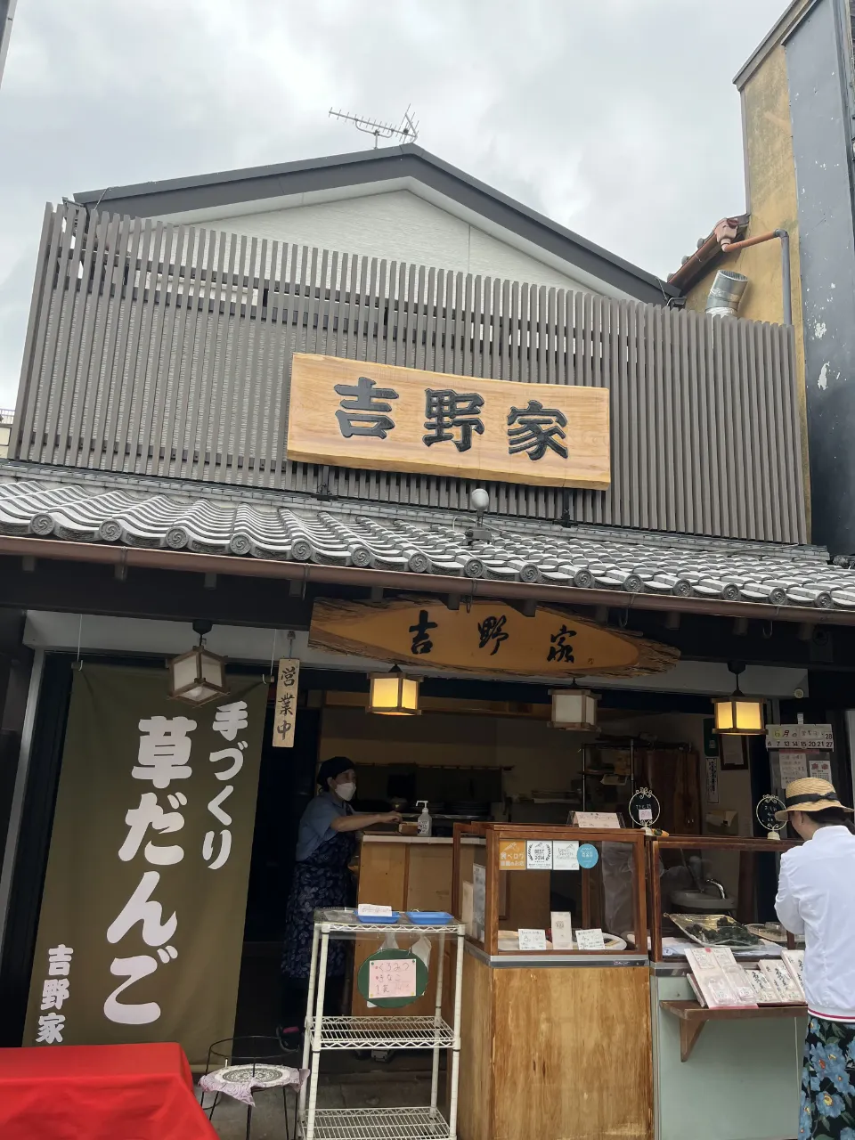 木招牌寫著吉野家的和菓子店，門口掛草だんご布簾，店員與客人在櫃前。