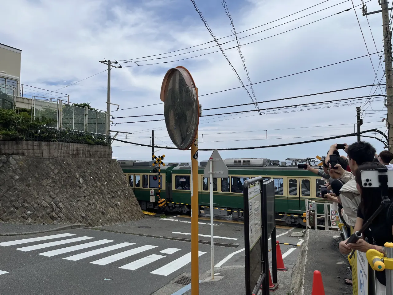江之電綠米色電車駛過平交道，欄杆邊一排遊客舉手機拍照