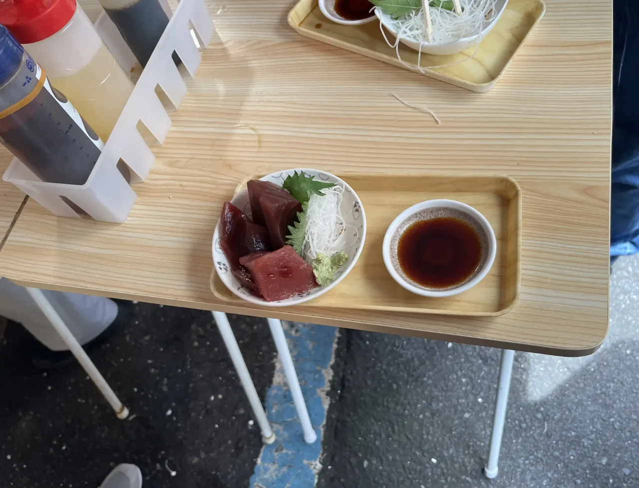 木のトレーにのったマグロの刺身の小鉢と、醤油の小皿。屋外のテーブル。